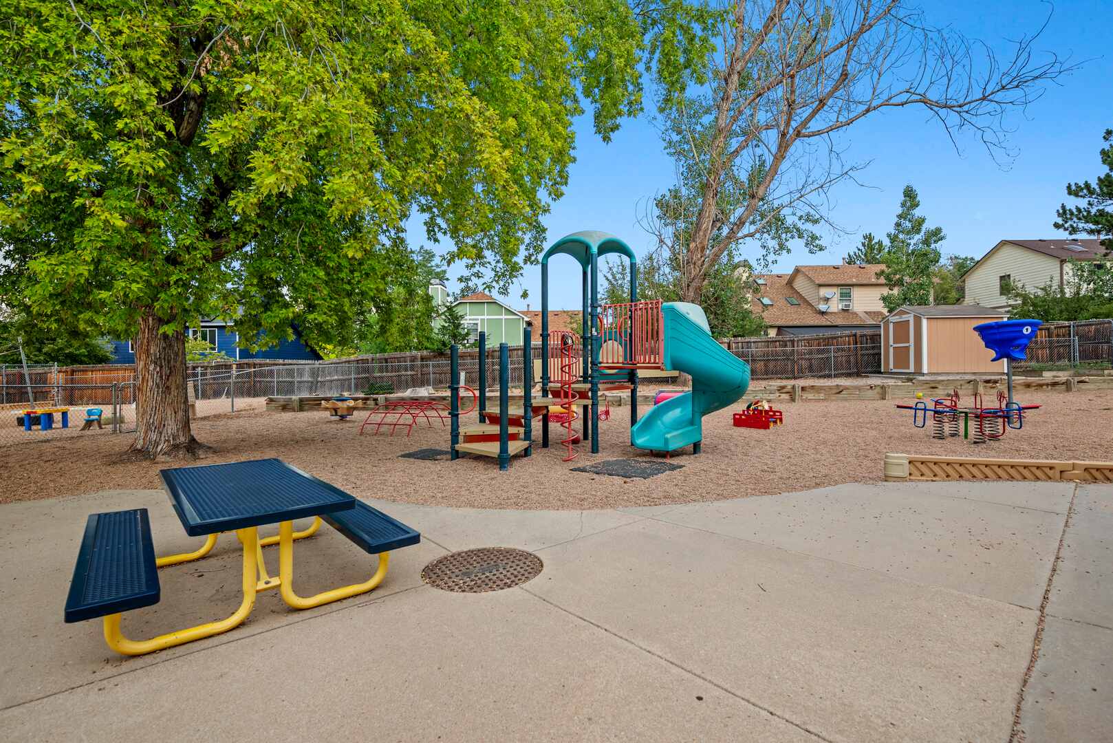 Outdoor playground with slides, climbing structure, and picnic table under shade trees