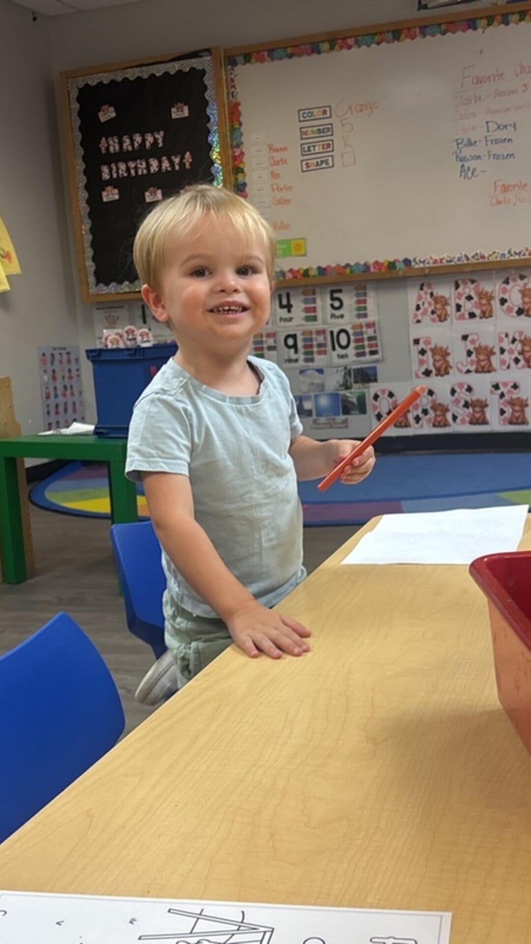 Smiling toddler standing at a classroom table holding a crayon