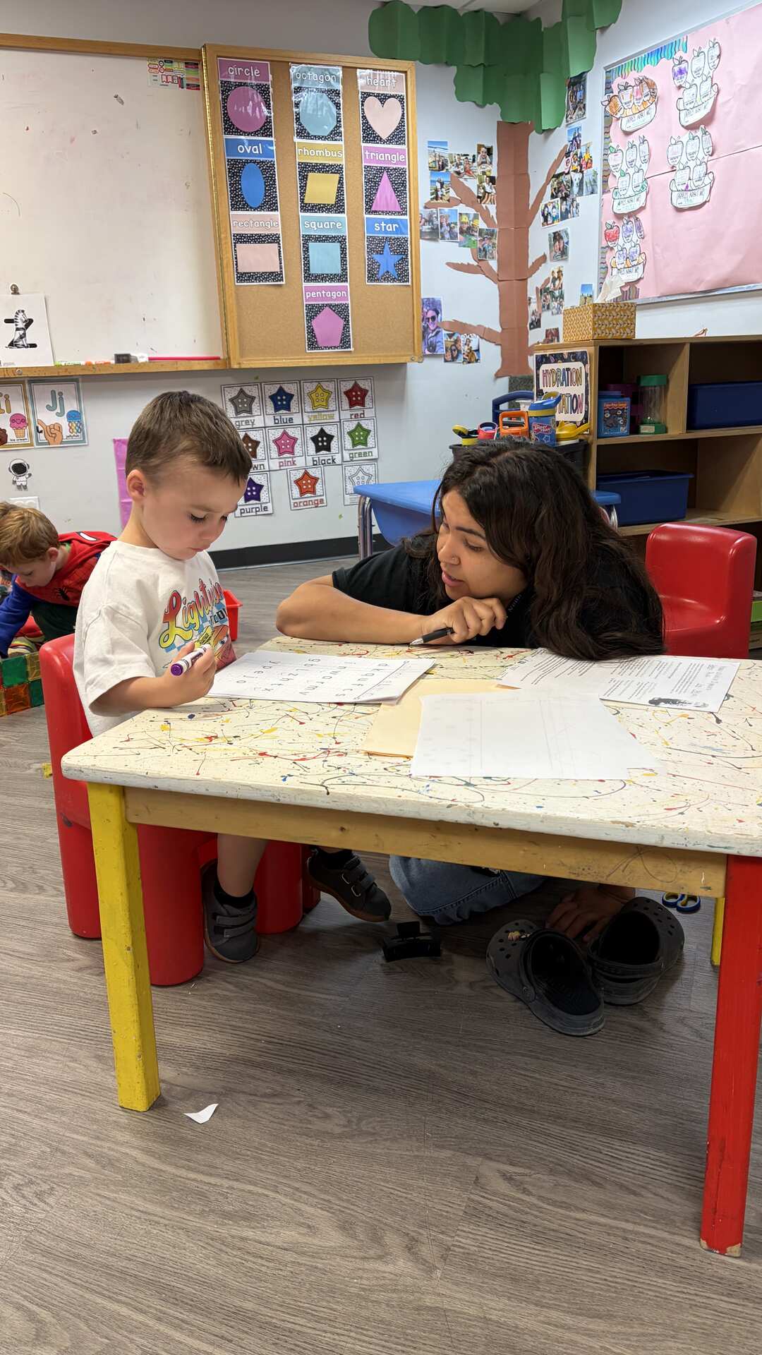Teacher helping a preschooler with a worksheet at a classroom table