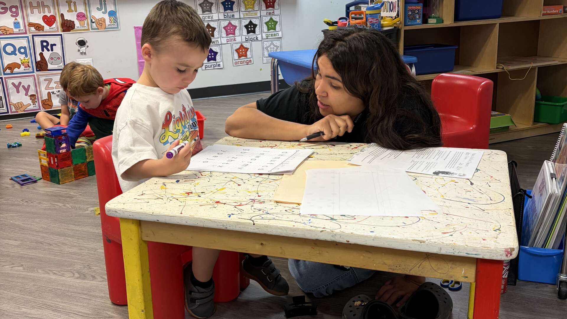 Teacher helping a preschooler with a worksheet at a classroom table